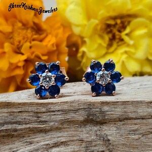 May include: Rose gold flower stud earrings with blue and clear gemstones. The earrings are set against a wooden surface with a blurred background of yellow and orange flowers. The earrings are from Shree Krishna Jewelers.