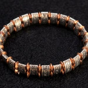 May include: A silver and copper-toned bracelet with a repeating pattern of etched silver cylinders and copper-colored beads. The bracelet is circular and rests on a black surface. The design is intricate and detailed, showcasing a blend of metallic textures.