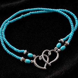 Peut inclure: Un collier de perles turquoise avec un pendentif double cœur. Le collier présente deux rangées de petites perles rondes et un fermoir en forme de cœur argenté. Les perles sont d'un bleu-vert vif.