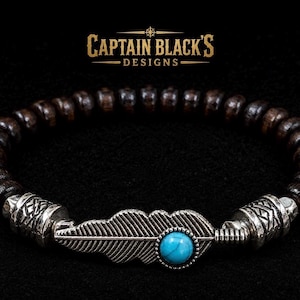 May include: A beaded bracelet featuring dark brown wooden beads and two silver-toned metal spacers with etched patterns. A central silver-toned feather charm is accented with a round, bright blue turquoise-coloured stone. Text reads CAPTAIN BLACK'S DESIGNS.