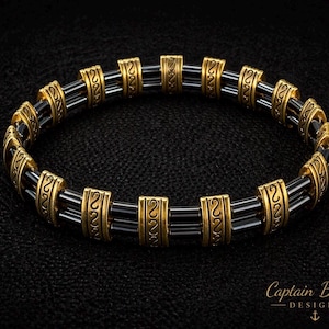 May include: A beaded bracelet with black and gold accents. The bracelet features black cylindrical beads separated by ornate gold-colored spacers. The spacers have a repeating design. The bracelet is displayed on a black surface. The text "Captain Black's Designs" is visible.