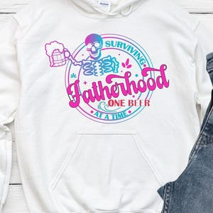 May include: White hoodie with a graphic that reads "Surviving Fatherhood One Beer At A Time." The design features a skeleton holding a beer mug, with a pink and blue color scheme. The hoodie has a front pocket and a drawstring hood.