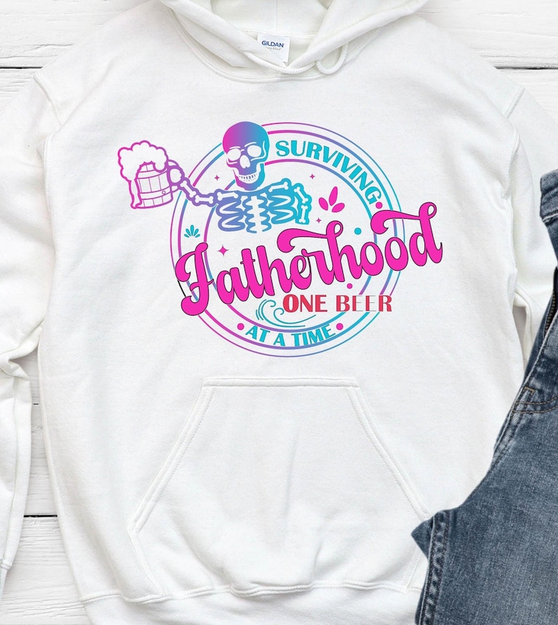 May include: White hoodie with a graphic that reads "Surviving Fatherhood One Beer At A Time." The design features a skeleton holding a beer mug, with a pink and blue color scheme. The hoodie has a front pocket and a drawstring hood.