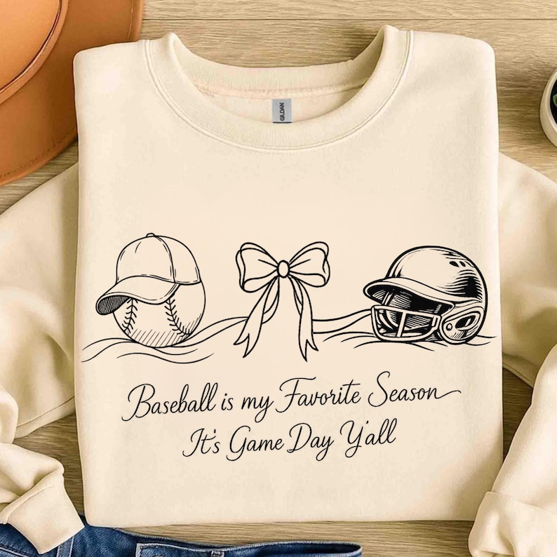 May include: Cream-colored sweatshirt with a black graphic of a baseball, a bow, and a baseball helmet. The text "Baseball is my Favorite Season It's Game Day Yall" is printed below the graphic.