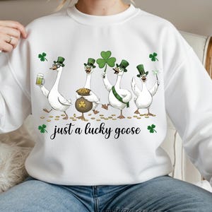 Puede incluir: Sudadera blanca con un dise&ntilde;o del D&iacute;a de San Patricio. El gr&aacute;fico muestra cuatro gansos de dibujos animados con sombreros verdes de copa y sosteniendo una cerveza, una olla de oro, un tr&eacute;bol y una bengala. El texto "just a lucky goose" est&aacute; debajo.