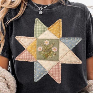 May include: Dark gray t-shirt featuring a star-shaped patchwork design. The star is composed of various fabric patterns, including gingham and floral prints, with a central square showcasing a floral bouquet. The design incorporates pastel colors.
