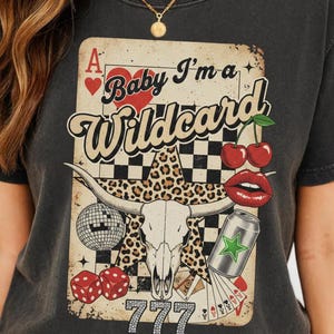 May include: Dark gray t-shirt featuring a graphic with a playing card design. The design includes a bull skull, cherries, dice, and the text "Baby I'm a Wildcard" and "777". The shirt has a vintage, distressed look.