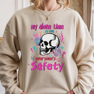 May include: Beige sweatshirt with a graphic design featuring a white skull, peace signs, butterflies, and lightning bolts. The text reads "my alone time is for everyone's safety" in pink and purple.