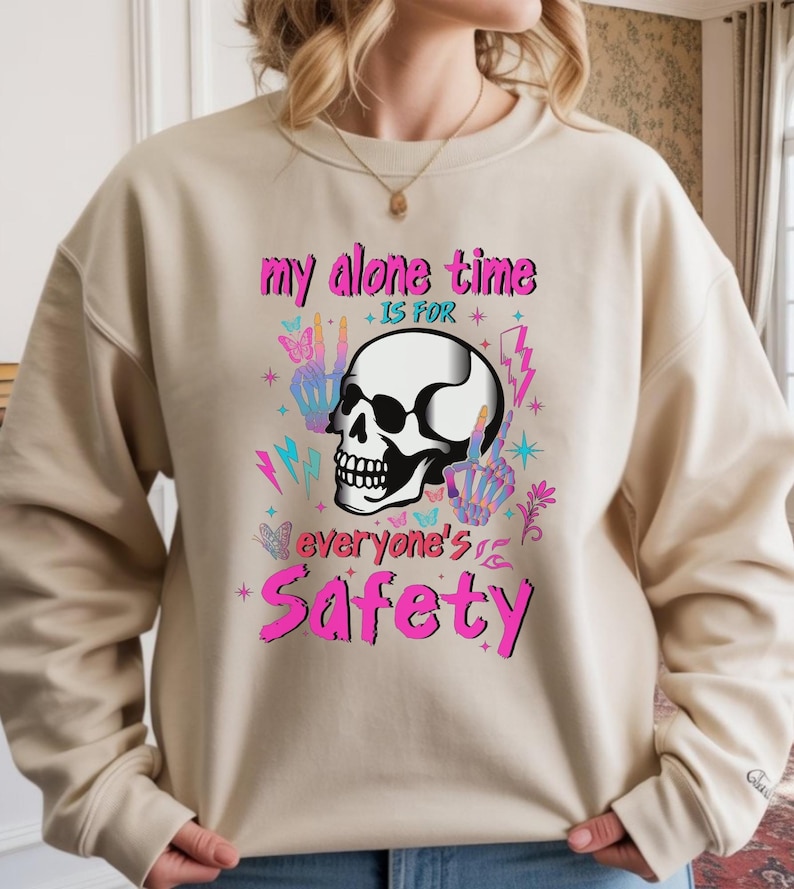 May include: Beige sweatshirt with a graphic design featuring a white skull, peace signs, butterflies, and lightning bolts. The text reads "my alone time is for everyone's safety" in pink and purple.