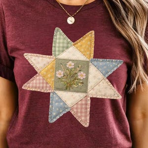 May include: Burgundy t-shirt featuring a patchwork star design. The star is composed of various patterned fabrics in pastel colors, with a central square showcasing embroidered flowers. The overall design has a vintage, handcrafted aesthetic.