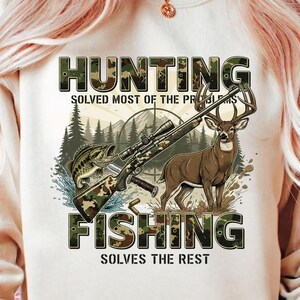 May include: Cream-colored graphic tee featuring the words "HUNTING" and "FISHING" in camouflage lettering. The design includes a rifle, a deer, a fish, and the text "SOLVES MOST OF THE PROBLEMS" and "SOLVES THE REST".