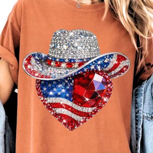 May include: A rhinestone-studded cowboy hat with red, white, and blue accents, and a heart-shaped pendant featuring the American flag and a large red gem. The design is set against a burnt orange t-shirt.