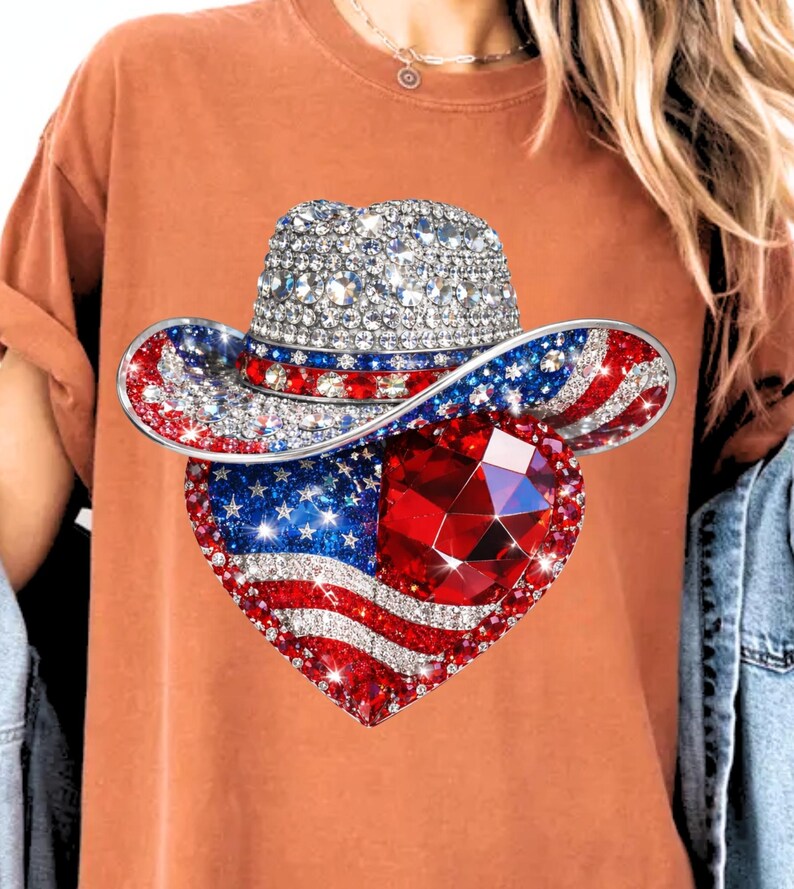 May include: A rhinestone-studded cowboy hat with red, white, and blue accents, and a heart-shaped pendant featuring the American flag and a large red gem. The design is set against a burnt orange t-shirt.