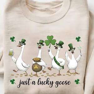 Puede incluir: Sudadera beige con un dise&ntilde;o caprichoso de cuatro gansos blancos vestidos con atuendos verdes del D&iacute;a de San Patricio. Un ganso sostiene una cerveza, otro una olla de oro y otro un gran tr&eacute;bol. El texto dice "just a lucky goose".