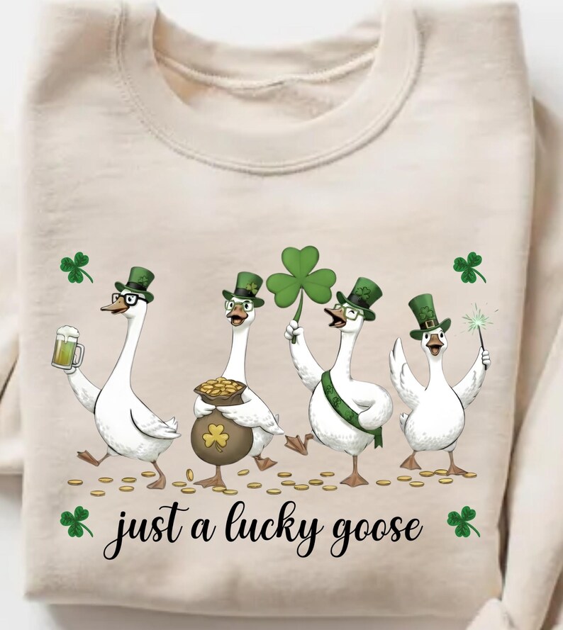 Puede incluir: Sudadera beige con un dise&ntilde;o caprichoso de cuatro gansos blancos vestidos con atuendos verdes del D&iacute;a de San Patricio. Un ganso sostiene una cerveza, otro una olla de oro y otro un gran tr&eacute;bol. El texto dice "just a lucky goose".