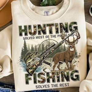 May include: Cream-colored sweatshirt with a hunting and fishing graphic. The design includes a rifle, a deer, a fish, and the text "HUNTING SOLVED MOST OF THE PROBLEMS FISHING SOLVES THE REST."