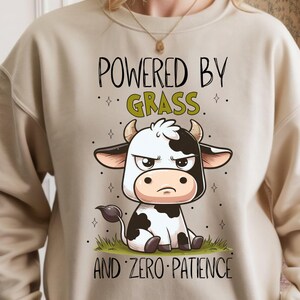 K&ouml;nnte beinhalten: Beiges Sweatshirt mit dem Text "POWERED BY GRASS" &uuml;ber einer Cartoon-Kuh-Illustration. Die Kuh ist schwarz-wei&szlig; mit einem w&uuml;tenden Gesichtsausdruck, und der Text "AND ZERO PATIENCE" steht unter der Kuh.