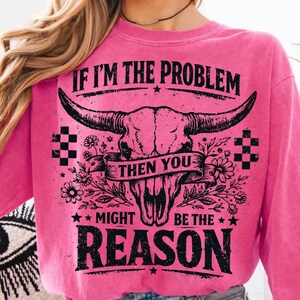 May include: A pink sweatshirt with a graphic of a bull skull and the text "IF I'M THE PROBLEM THEN YOU MIGHT BE THE REASON" in black. The design includes floral and checkered elements.
