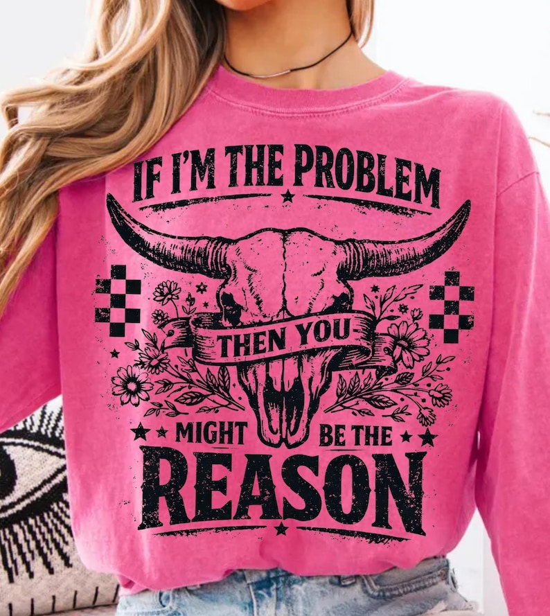 May include: A pink sweatshirt with a graphic of a bull skull and the text "IF I'M THE PROBLEM THEN YOU MIGHT BE THE REASON" in black. The design includes floral and checkered elements.