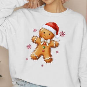 May include: White sweatshirt featuring a gingerbread man design. The gingerbread man wears a red Santa hat and a white bow tie. Red snowflakes surround the gingerbread man. The design is centered on the front of the sweatshirt.
