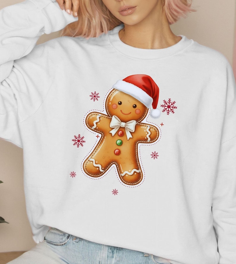 May include: White sweatshirt featuring a gingerbread man design. The gingerbread man wears a red Santa hat and a white bow tie. Red snowflakes surround the gingerbread man. The design is centered on the front of the sweatshirt.