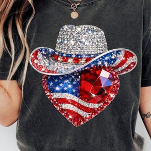 May include: Dark gray t-shirt featuring a sparkling cowboy hat and a heart-shaped design. The hat and heart are adorned with red, white, and blue rhinestones, creating a patriotic theme. The heart has a large, faceted red gem.