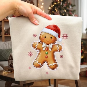 May include: A beige Christmas-themed pillow featuring a gingerbread man wearing a Santa hat and a white bow tie. Red and white snowflakes surround the gingerbread man. The pillow is held up by a hand.