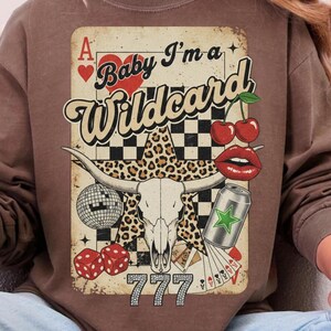 May include: Brown sweatshirt with a graphic design featuring a playing card, a bull skull, cherries, dice, and the text "Baby I'm a Wildcard". The design includes a leopard print star and the number "777".