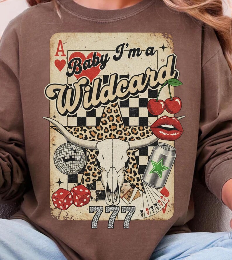 May include: Brown sweatshirt with a graphic design featuring a playing card, a bull skull, cherries, dice, and the text "Baby I'm a Wildcard". The design includes a leopard print star and the number "777".