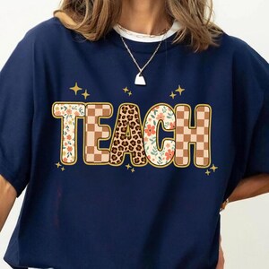 May include: Navy blue sweatshirt with the word "TEACH" in a decorative font. The letters are adorned with floral, leopard print, and checkered patterns, with gold star accents.