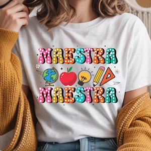 May include: White t-shirt with the word "MAESTRA" in colorful, bubble letters. The shirt features school-themed graphics, including a globe, apple, lightbulb, pencil, and ruler. The design is in a retro style.