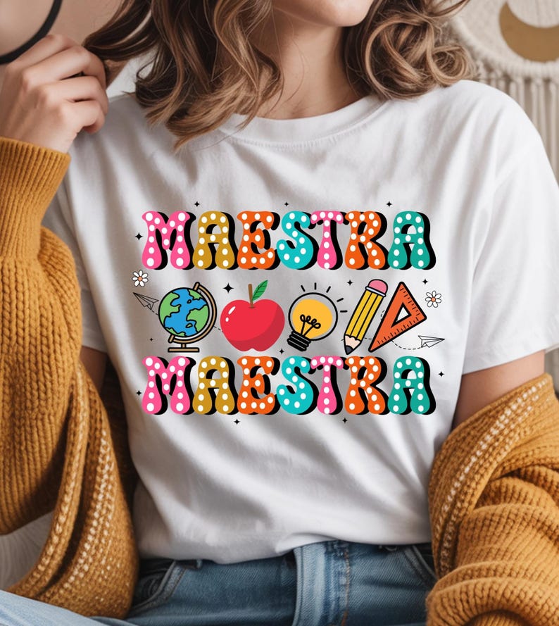 May include: White t-shirt with the word "MAESTRA" in colorful, bubble letters. The shirt features school-themed graphics, including a globe, apple, lightbulb, pencil, and ruler. The design is in a retro style.