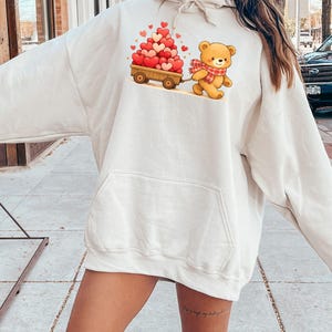 May include: Cream-coloured hoodie with a cartoon bear pulling a wooden cart filled with heart-shaped balloons. The bear wears a red and white scarf. The hoodie has a front pocket and a drawstring hood.