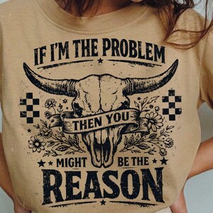 May include: Tan-colored t-shirt featuring a graphic of a bull skull with large horns, floral accents, and the text "IF I'M THE PROBLEM THEN YOU MIGHT BE THE REASON" in black. The design includes stars and checkered patterns.