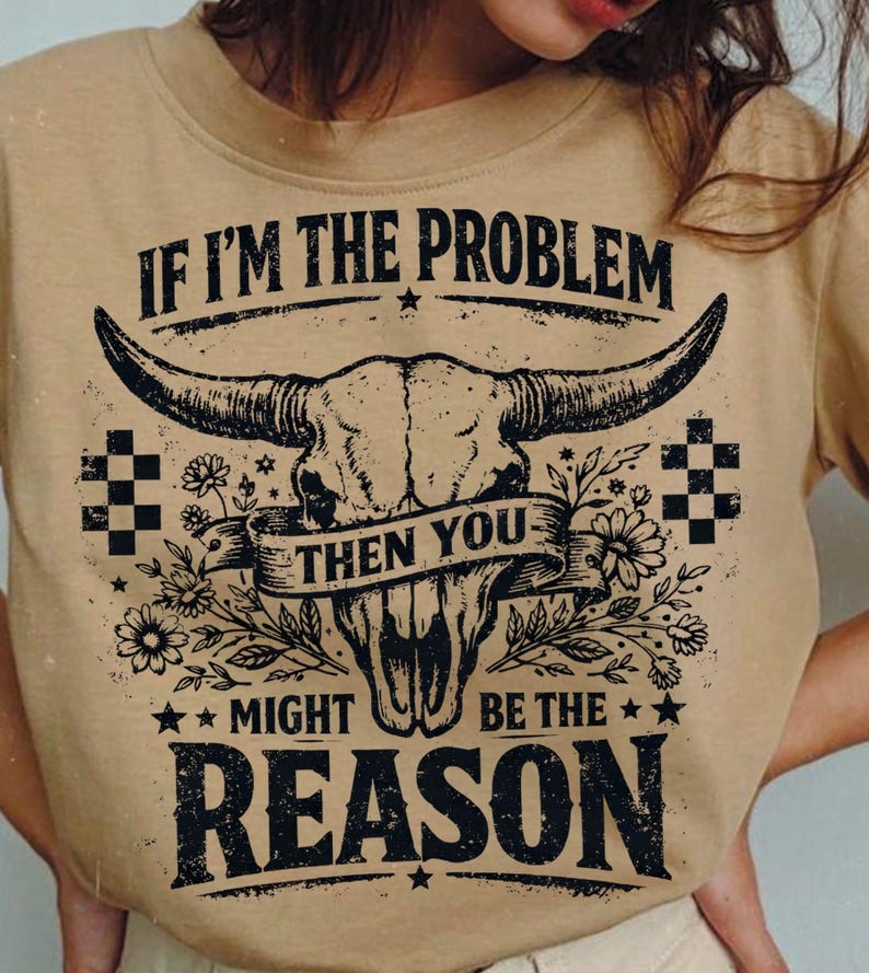 May include: Tan-colored t-shirt featuring a graphic of a bull skull with large horns, floral accents, and the text "IF I'M THE PROBLEM THEN YOU MIGHT BE THE REASON" in black. The design includes stars and checkered patterns.