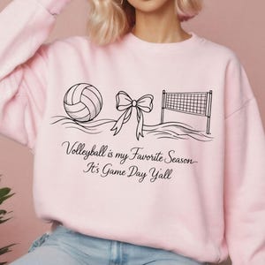 May include: A light pink sweatshirt with a black graphic of a volleyball, a bow, and a volleyball net. The text "Volleyball is my Favorite Season It's Game Day Y'all" is printed below the graphic.
