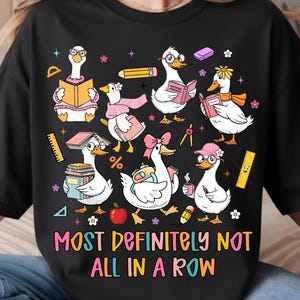 May include: Black sweatshirt featuring a whimsical design of cartoon geese wearing glasses and reading books, with pencils, and flowers. The text "MOST DEFINITELY NOT ALL IN A ROW" is displayed in colorful letters.
