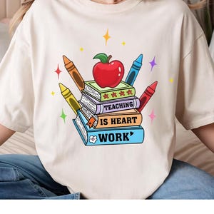 May include: Cream-colored t-shirt featuring a colorful graphic design. The design includes a stack of books with the words "TEACHING IS HEART WORK" and a red apple on top. Crayons in various colors surround the books, with star accents.