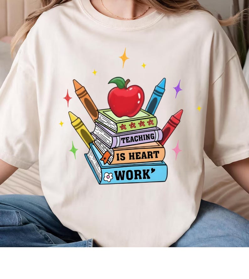May include: Cream-colored t-shirt featuring a colorful graphic design. The design includes a stack of books with the words "TEACHING IS HEART WORK" and a red apple on top. Crayons in various colors surround the books, with star accents.