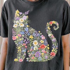 May include: Black t-shirt featuring a floral cat design. The cat silhouette is filled with colorful flowers, including daisies, poppies, and wildflowers in shades of pink, yellow, blue, and purple. The design is centered on the front of the shirt.