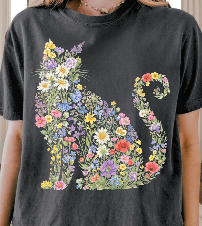 May include: Black t-shirt featuring a floral cat design. The cat silhouette is filled with colorful flowers, including daisies, poppies, and wildflowers in shades of pink, yellow, blue, and purple. The design is centered on the front of the shirt.