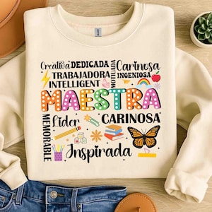 May include: Cream-colored sweatshirt with the word "MAESTRA" in colorful, dotted letters. Surrounding the word are other words like "Creativa," "Dedicada," and "Cari&ntilde;osa," along with illustrations of a butterfly, pencil, and books.