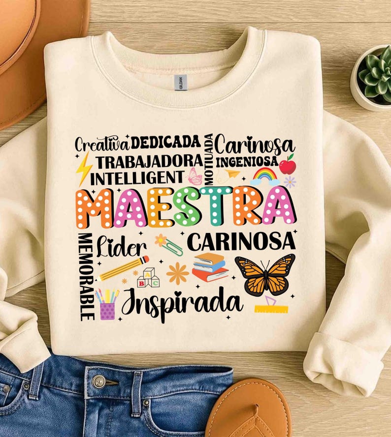 May include: Cream-colored sweatshirt with the word "MAESTRA" in colorful, dotted letters. Surrounding the word are other words like "Creativa," "Dedicada," and "Cari&ntilde;osa," along with illustrations of a butterfly, pencil, and books.