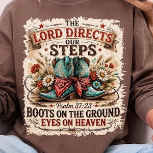 May include: Brown sweatshirt with a graphic featuring turquoise cowboy boots, a red bandana, and floral accents. Text includes "The Lord Directs Our Steps," "Psalm 37:23," and "Boots on the Ground Eyes on Heaven."