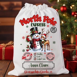 May include: White drawstring gift bag with a festive design. The bag features the text "North Pole Express" in red, along with illustrations of a snowman and a reindeer. Includes the name "Anna Claire" and the message "Do not open before December 25".