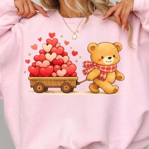 May include: Pink sweatshirt with a cartoon bear pulling a cart of hearts. The bear wears a red and white checked scarf. The cart is filled with red and pink heart-shaped balloons. A cute design for Valentine's Day.