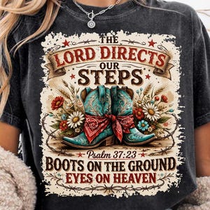 May include: Black t-shirt featuring a graphic with turquoise cowboy boots, a red bandana, and floral accents. The text reads "The Lord Directs Our Steps" and "Boots on the Ground, Eyes on Heaven."