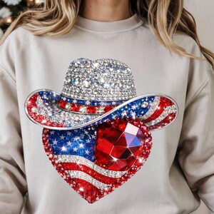 May include: A beige sweatshirt featuring a sparkling cowboy hat and a heart-shaped design. The hat is embellished with clear rhinestones and the brim is decorated with red, white, and blue accents. The heart showcases the American flag and a large red gem.