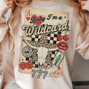 May include: Cream-colored sweatshirt featuring a graphic of a playing card with the text "Baby I'm a Wildcard." The design includes a bull skull, cherries, dice, and a disco ball. The card has a vintage, distressed look.