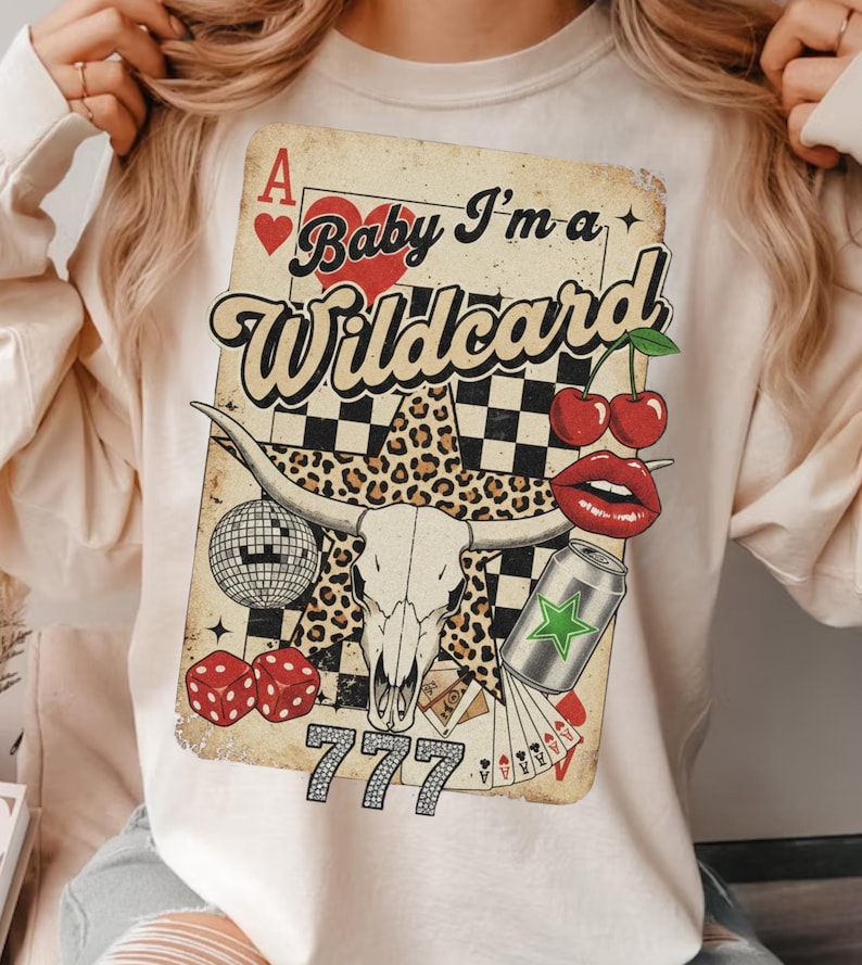 May include: Cream-colored sweatshirt featuring a graphic of a playing card with the text "Baby I'm a Wildcard." The design includes a bull skull, cherries, dice, and a disco ball. The card has a vintage, distressed look.